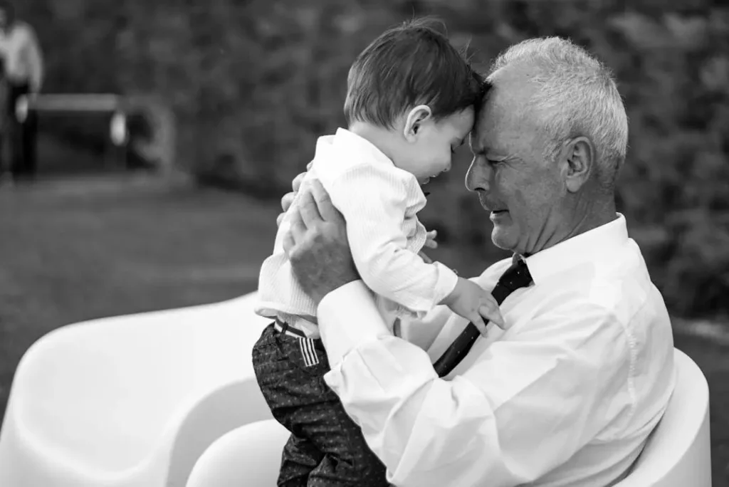 A black-and-white photo of an elderly man tenderly holding a young child. The two are facing each other closely, gently touching their foreheads. The man is wearing a white shirt and tie, while the child is dressed in a long-sleeve shirt and pants. The background appears to be outdoors, with blurred greenery visible.